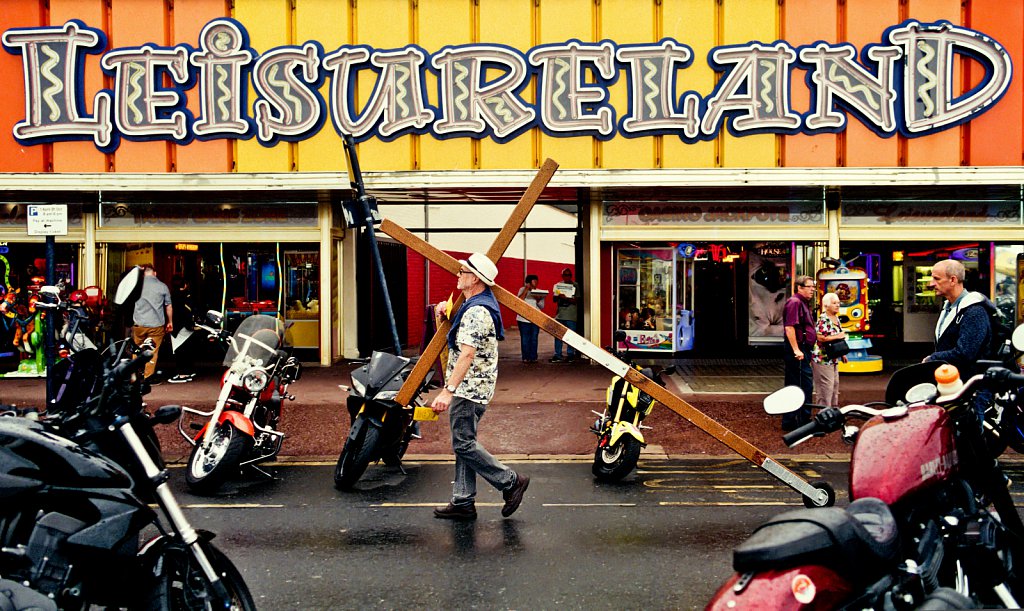 An evangelical preacher hauls a crucifix past the Leiesureland amusement arcade.